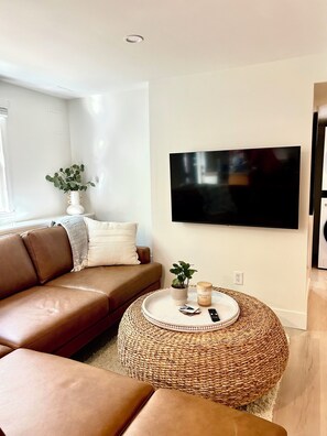 A living room with a brown leather sofa, a round woven ottoman, a flat-screen TV mounted on the wall, and a small plant on the sofa. - Charming 2-bedroom apartment in amazing Dennis (Dennis)