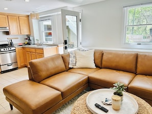A modern kitchen with wooden cabinets, a stainless steel stove, and a dining area with a brown leather sofa and a round coffee table. - Charming 2-bedroom apartment in amazing Dennis (Dennis)