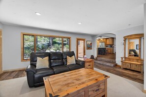 Ein Wohnzimmer mit schwarzem Ledersofa, einem Holztisch und großen Fenstern mit Blick auf Bäume.