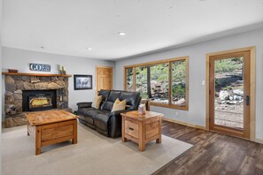 Ein Wohnzimmer mit Kamin aus Stein, einem Ledersofa, einem Holztisch und einem kleinen Beistelltisch. Große Fenster bieten Blick auf eine felsige Landschaft.