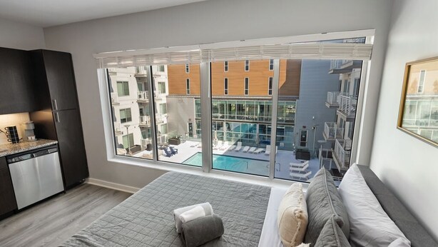 Ett modernt hotellrum med ett stort fönster, en säng, ett litet pentry och utsikt över en pool och byggnader.