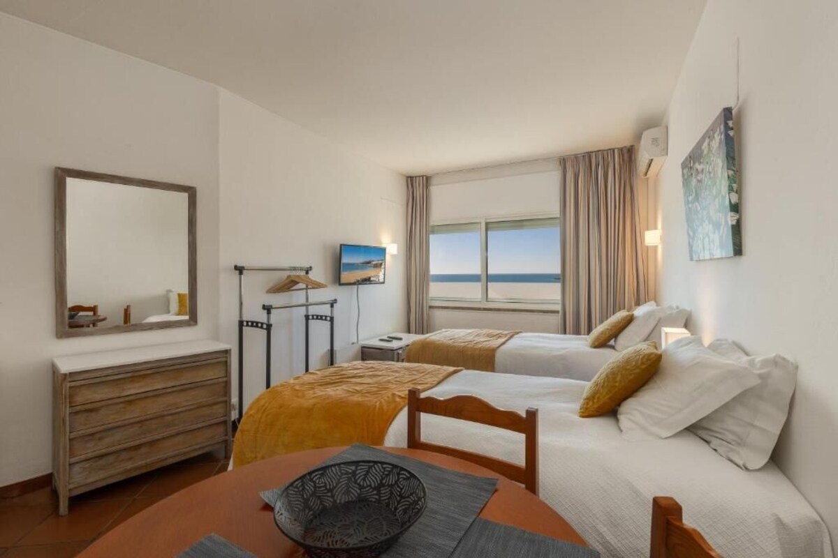 Ein Hotelzimmer mit zwei Betten, einem Esstisch, einer Kommode, einem Gemälde und Meerblick.