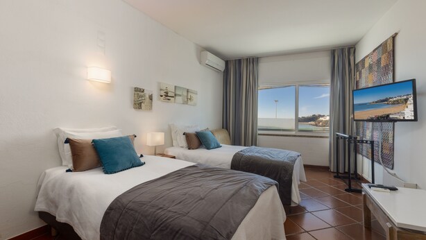 A hotel room with two beds, a TV, artwork on the wall, and a window with a view. - APARTAMENTOS PESCADORES (Albufeira)