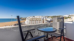A balcony with a glass table and chairs, overlooking a beach and a coastal town. - APARTAMENTOS PESCADORES (Albufeira)
