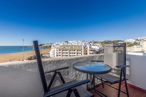 A balcony with a glass table and chairs, overlooking a beach and a coastal town. - APARTAMENTOS PESCADORES (Albufeira)