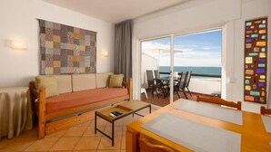 A room with a sofa, a table, a view of the sea, and a large window. - APARTAMENTOS PESCADORES (Albufeira)