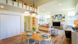 A spacious living area with a dining table set for four, a sofa, a TV, and a staircase leading to an upper level. - Cottage by the Sea (Whitethorn)