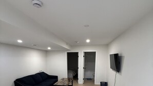 Ett rymligt vardagsrum med en soffa, ett soffbord, en platt-TV som är monterad på väggen och en dörr som leder till ett annat rum.