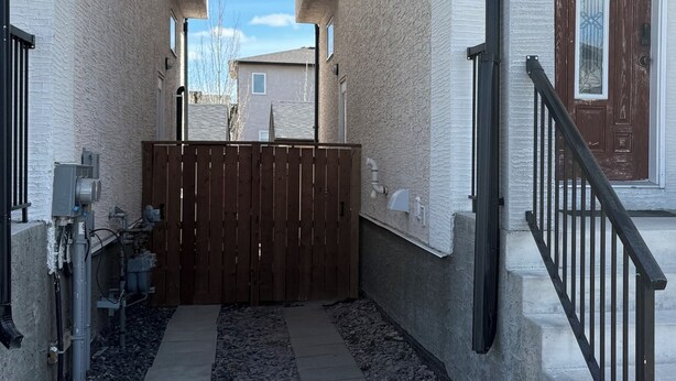 En smal gångväg leder fram till en grindad entré, och på var sin sida om den står två byggnader, medan himlen ovanför är klar och blå.