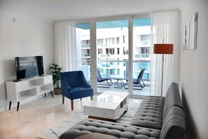 A modern living room with a sofa, a TV, a blue chair, a white coffee table, and a balcony with outdoor seating. - 2 X 2 Bedroom Suites Partial OceanView, beachaccess adjacent rooms C D E F units (Miami Beach)