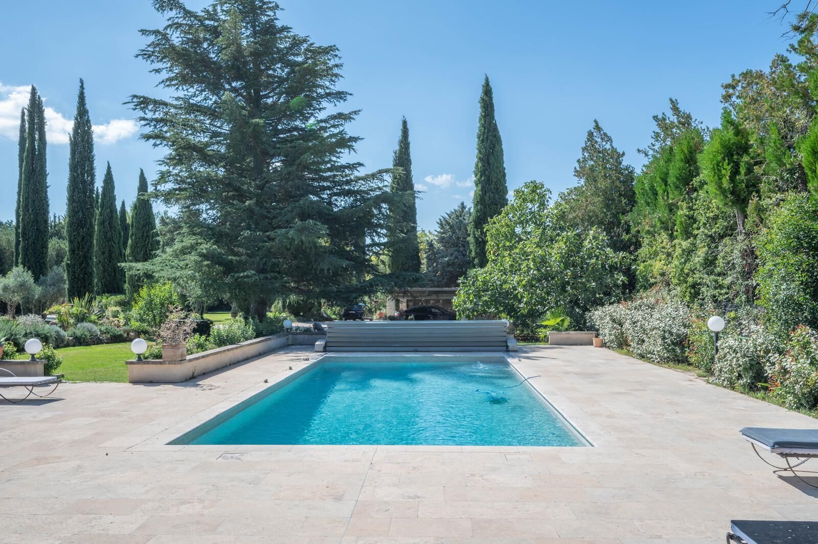 A rectangular swimming pool with a stone deck, surrounded by tall trees and a well-maintained garden.