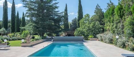 A rectangular swimming pool with a stone deck, surrounded by tall trees and a well-maintained garden.