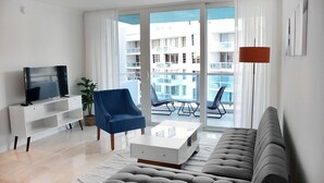 Ein modernes Wohnzimmer mit einer Couch, einem Fernseher, einem blauen Sessel und einem Balkon mit Sitzgelegenheiten.