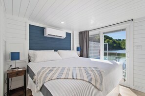 A bedroom with a large bed, a nightstand, a window with a view of greenery, and an air conditioning unit.