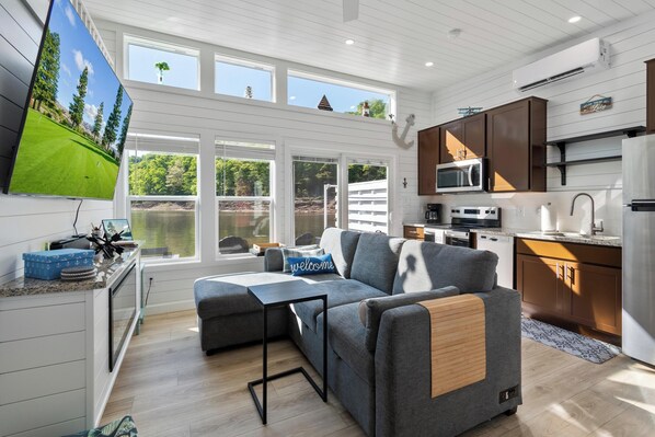 A modern kitchen with a flat-screen TV, wooden cabinets, and a large window overlooking a deck.