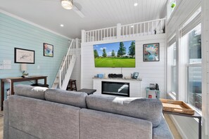 A living room with a grey sofa, a TV mounted on the wall, a fireplace, and a staircase leading to an upper level.