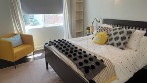 Ein Schlafzimmer mit einem Bett, einem Sessel, einem Nachttisch, einer Lampe, einer Pendelleuchte und einem Fenster mit Vorhängen.