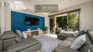 A modern living room with grey sofas, a flat-screen TV, and a sliding glass door leading to a garden. - Tides Edge in Capel Sound (Capel Sound)