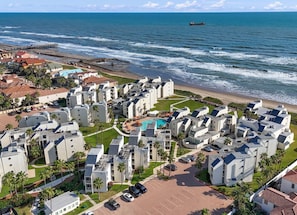 A coastal residential area with modern buildings, a central swimming pool, and a pier in the background.