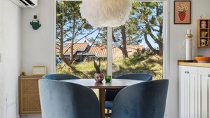 Un coin repas avec une table et des chaises, un luminaire suspendu et une vue sur une maison et des arbres, visible à travers une fenêtre.