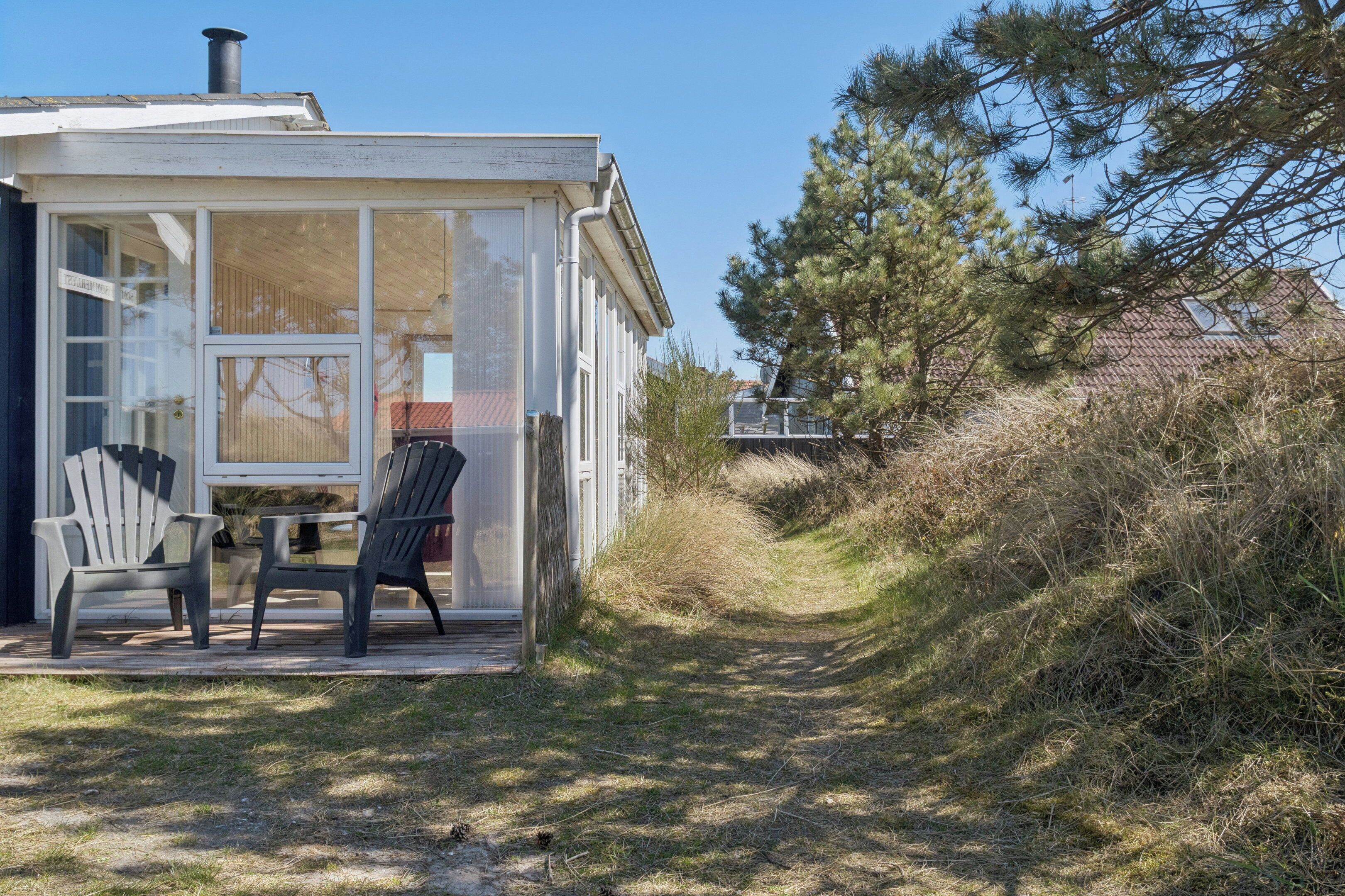 Una piccola casa a un piano con una porta e finestre in vetro, due poltrone Adirondack all'esterno e un vialetto che conduce a una casa sullo sfondo.