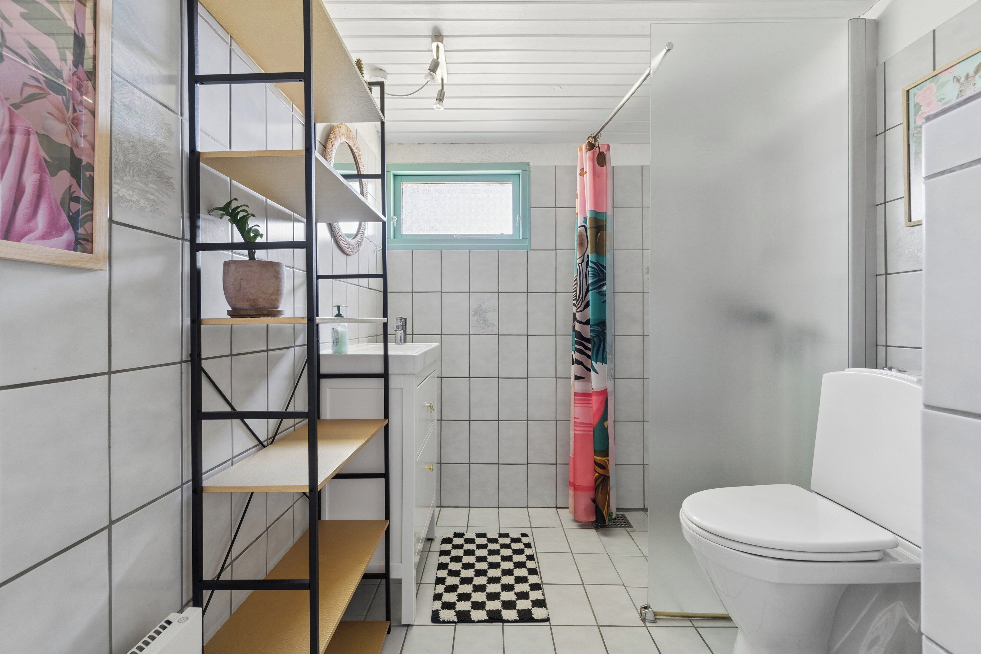 A bathroom with a toilet, a sink, a shower with a colorful curtain, a window, and a shelf with a potted plant.