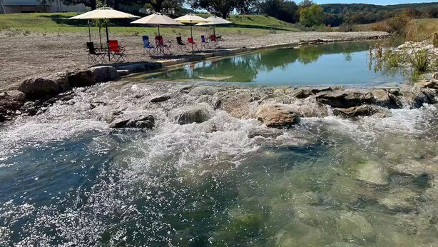 A river with clear water, rocks, and a row of beach chairs and umbrellas. - Hill Country Family & Pickleball Retreat (Ingram)