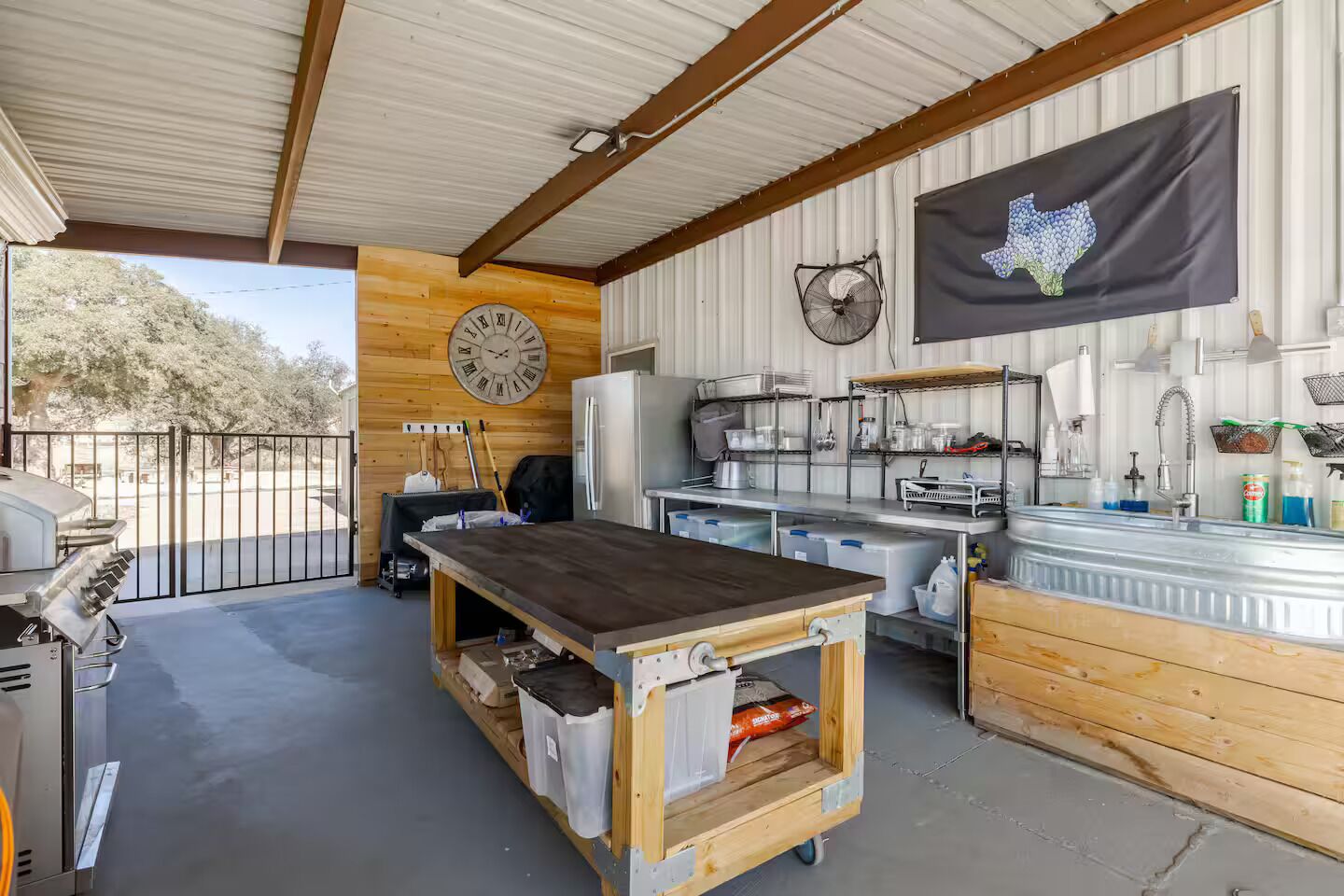 Un área de cocina al aire libre cubierta, con un reloj grande, una bandera de Texas y vistas a árboles.