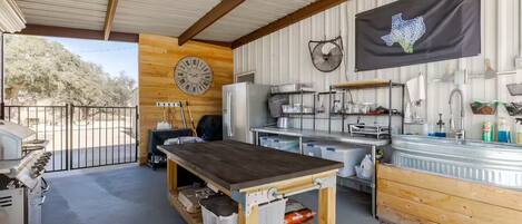 Un área de cocina al aire libre cubierta, con un reloj grande, una bandera de Texas y vistas a árboles.