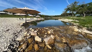 A clear outdoor area with a small pond, picnic tables, and umbrellas. - Hill Country Family & Pickleball Retreat (Ingram)