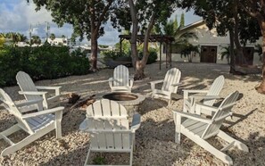 A set of Adirondack chairs arranged around a fire pit in a gravel area with trees and a building in the background. - Key Largo 2BR Retreat | Pool & Boat Ramp (Key Largo)
