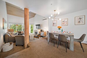 Uma sala de estar moderna com lareira, uma área de jantar com mesa posta para quatro pessoas e uma grande janela com vista para o verde.