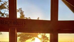 Blick aus einem Fenster, ein Schild mit der Aufschrift 'BEACH HOUSE' und ein Sonnenuntergang über dem Wasser.