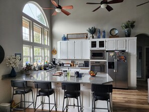 Eine moderne Küche mit weißen Schränken, einer großen Kochinsel mit Barhocker und einem Deckenventilator, mit Blick ins Freie durch große Fenster.