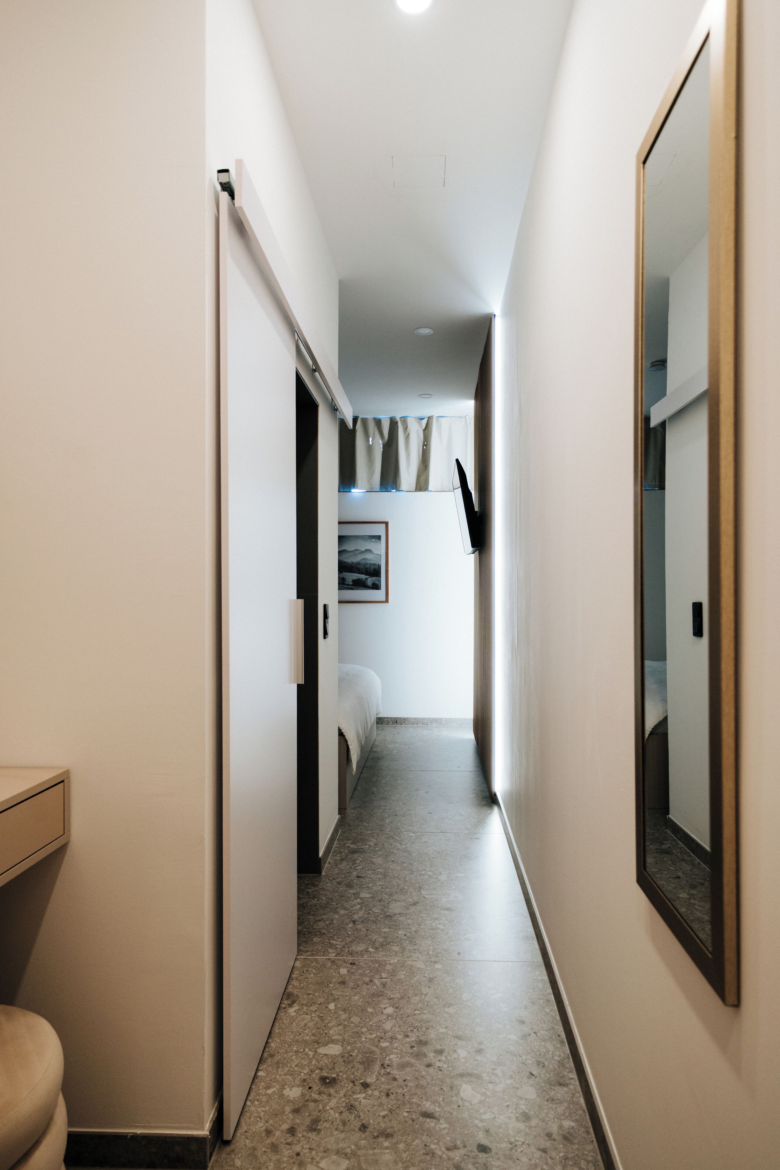 Een smalle gang met een bed, een spiegel en een deur naar een andere kamer.