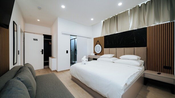 A modern hotel room with a large bed, a sofa, a bedside table, a mirror, and a wardrobe.