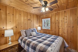 Ein Schlafzimmer mit Holzwänden, einem Bett, zwei Nachttischlampen, einem Deckenventilator und einem Fenster mit Blick auf einen Wald.
