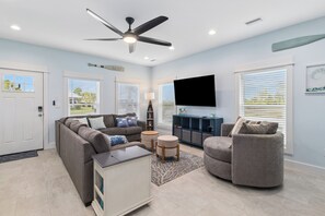 A living room with a sofa, armchair, ottoman, TV, ceiling fan, and a view of the outdoors.