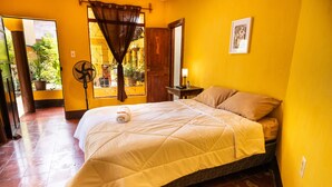 Una habitación con cama, una mesita de noche con lámpara, un ventilador y una ventana con cortinas.