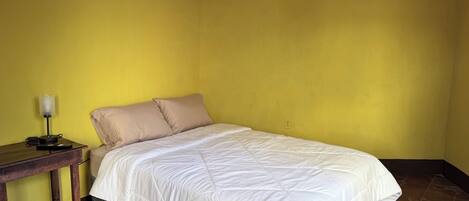Una cama individual con edredón blanco en una habitación con paredes amarillas y una mesita de noche de madera con lámpara.