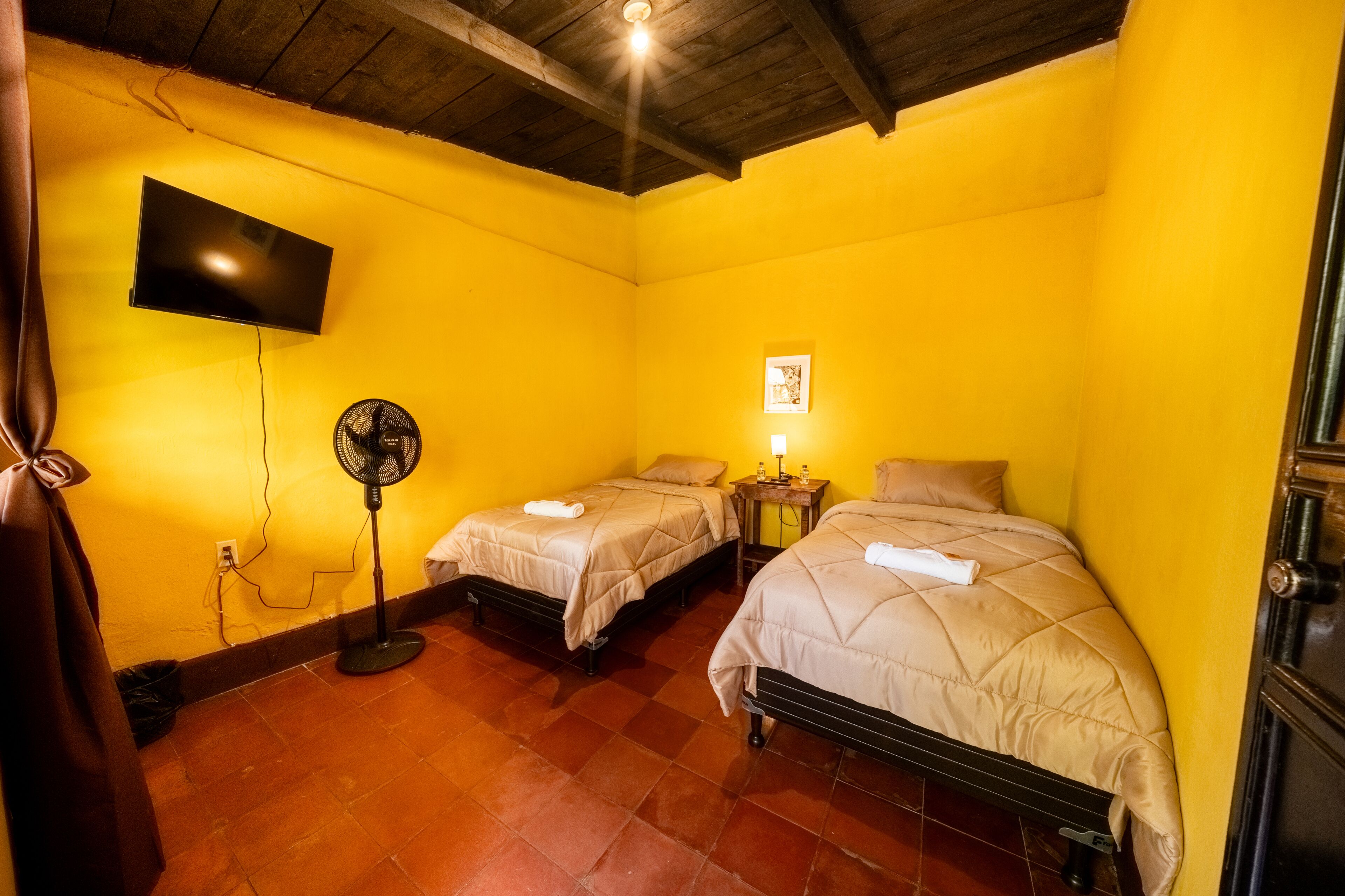 Una habitación con dos camas, un televisor montado en la pared, un ventilador y una mesita con una lámpara.