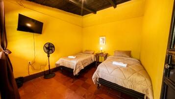Una habitación con dos camas, un televisor montado en la pared, un ventilador y una mesita con una lámpara.