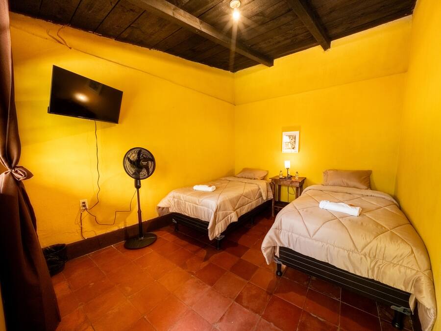 Una habitación con dos camas, un televisor montado en la pared, un ventilador y una mesita con una lámpara.