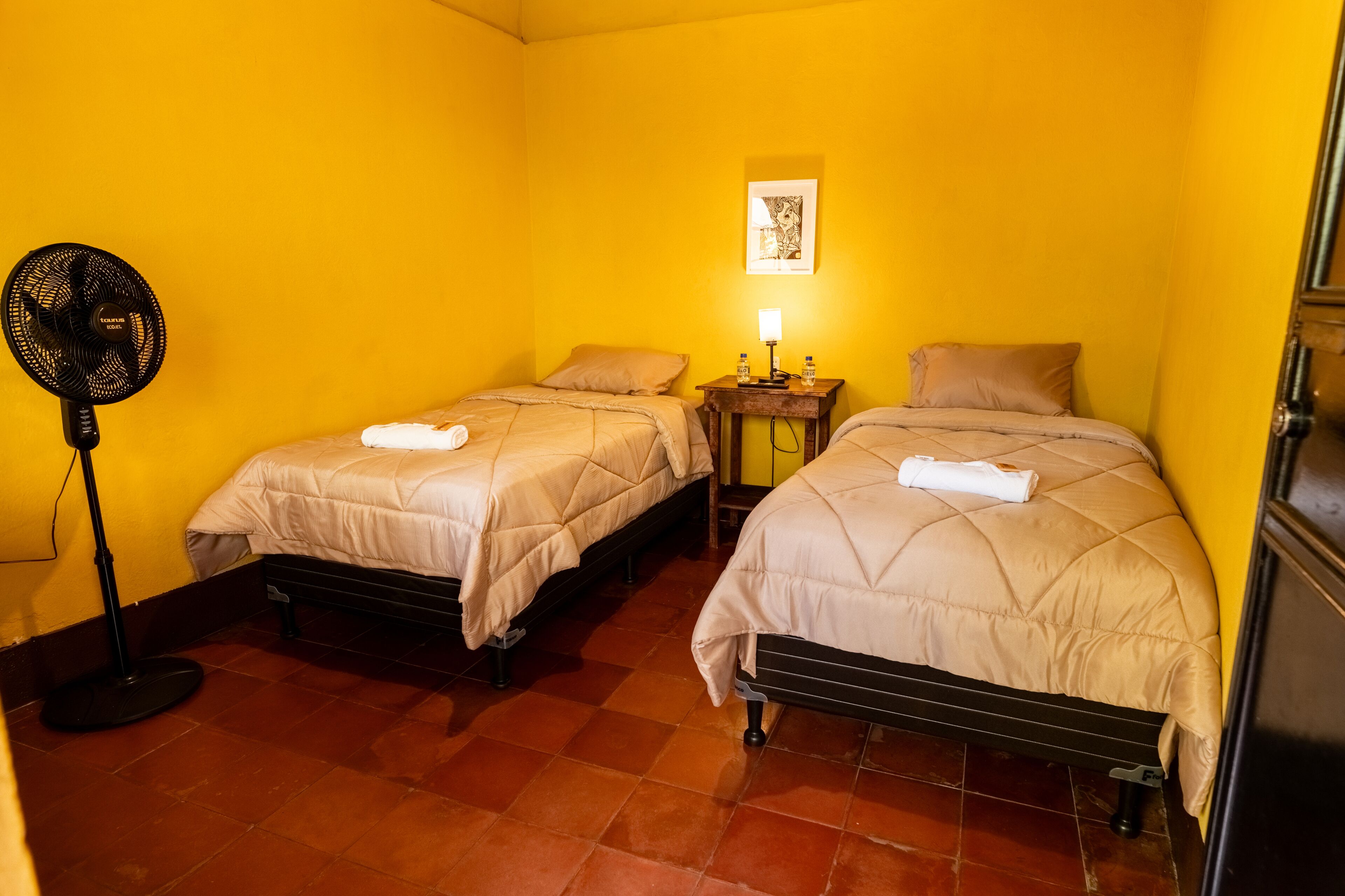 A room with two beds, a standing fan, a nightstand with a lamp, and a framed picture on the wall.