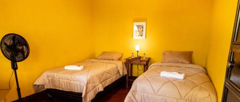 A room with two beds, a standing fan, a nightstand with a lamp, and a framed picture on the wall.