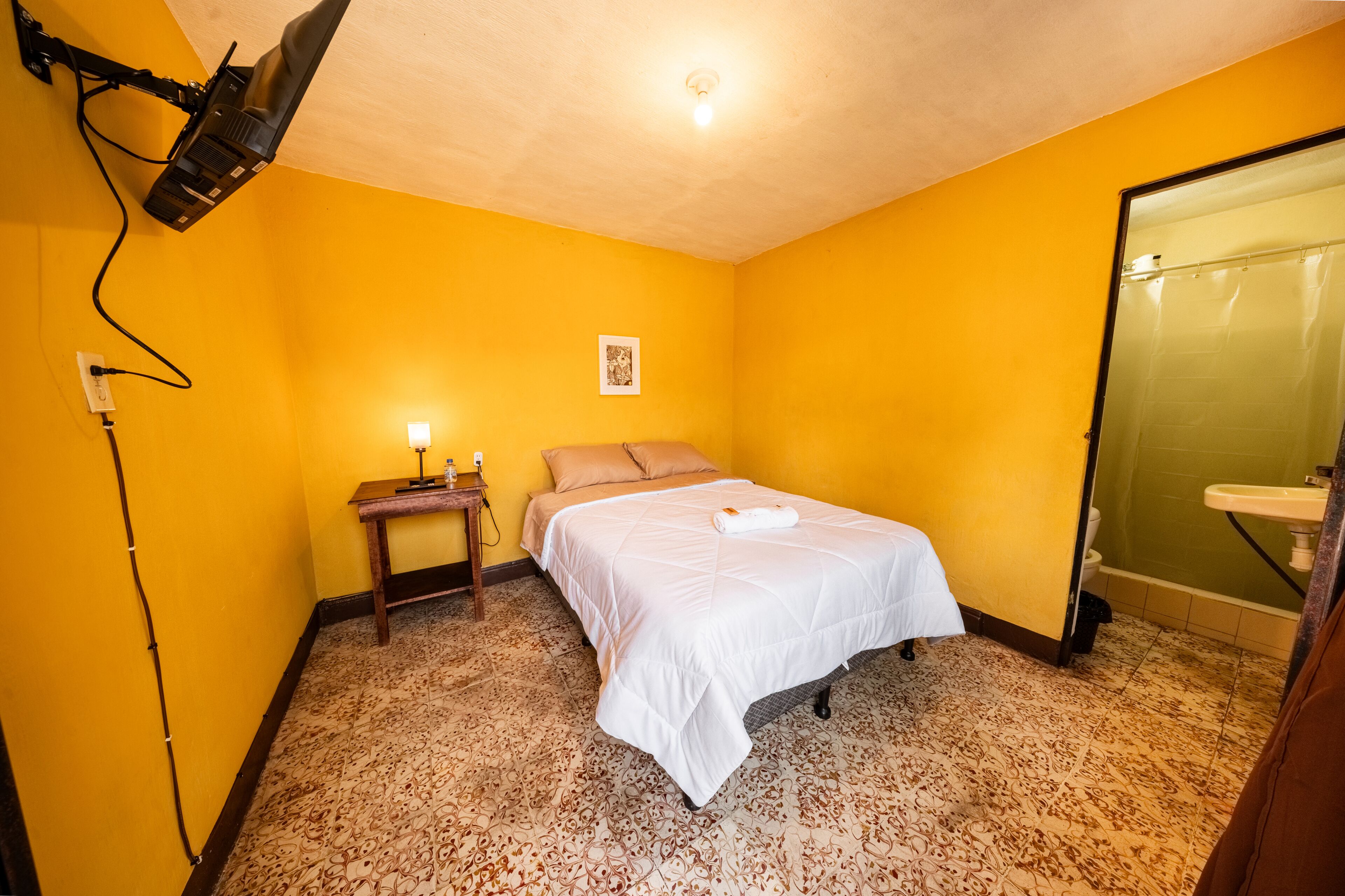 Una habitación con cama, mesita de noche, lámpara de pared y baño con lavamanos y ducha.