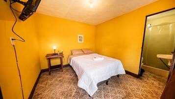 Una habitación con cama, mesita de noche, lámpara de pared y baño con lavamanos y ducha.