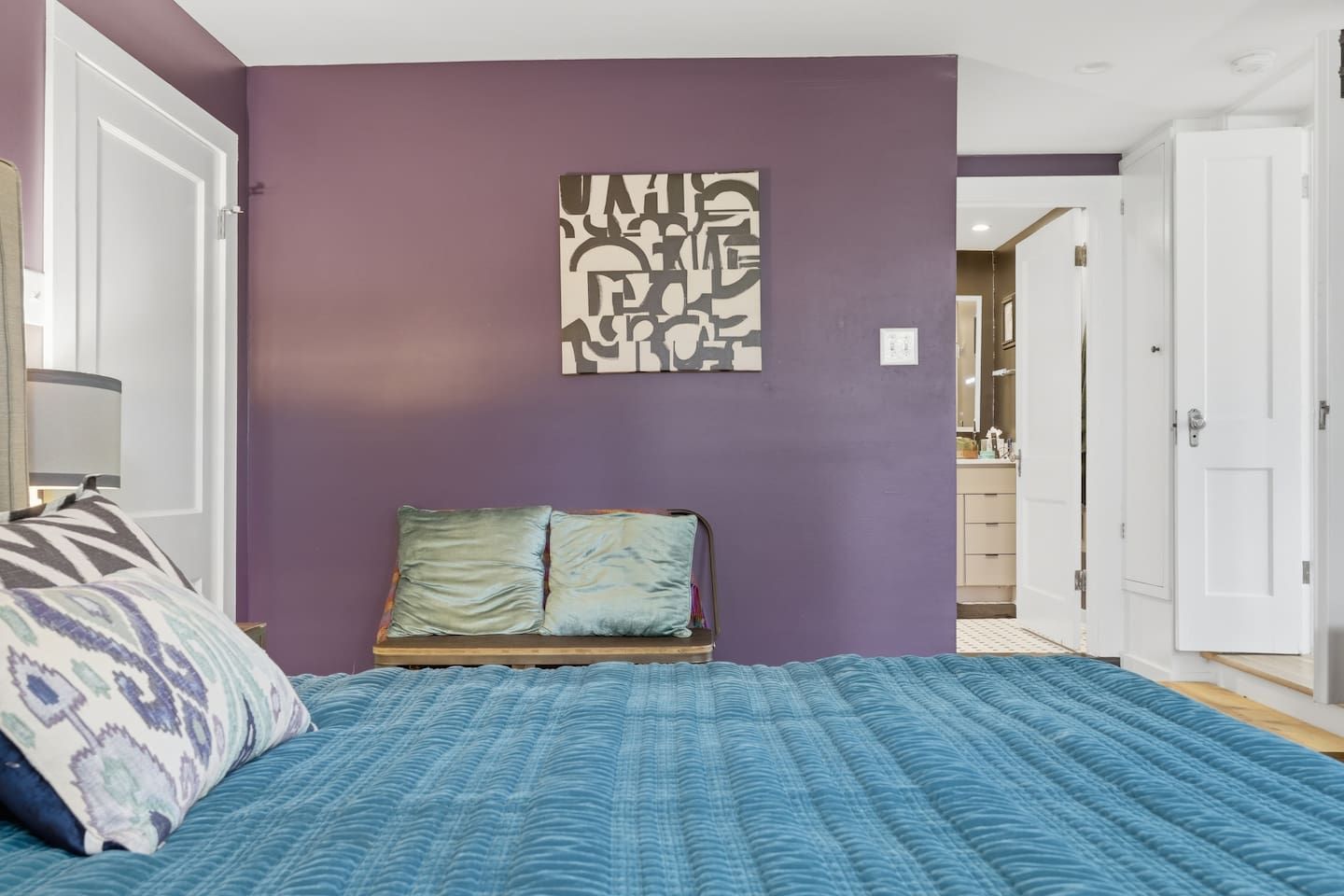 A bedroom with a bed, a headboard, a wall art piece, and a door leading to a bathroom.