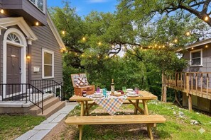 Un coin pique-nique aménagé dans un jardin, avec une table, des bancs et des guirlandes lumineuses, le tout entouré d’arbres et adossé à une maison.