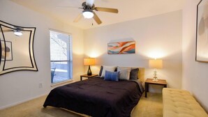 A bedroom with a bed, two bedside lamps, a ceiling fan, a window with blinds, and a wall-mounted abstract painting.